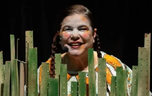 Regina França em "A Chave do Labirinto". A atriz está atrás de diversas réguas tingidas de verde, representando um canavial. Seu rosto foi pintado com maquiagem branca, batom vermelho e blush alaranjado com pintinhas; seu cabelo está preso em duas tranças