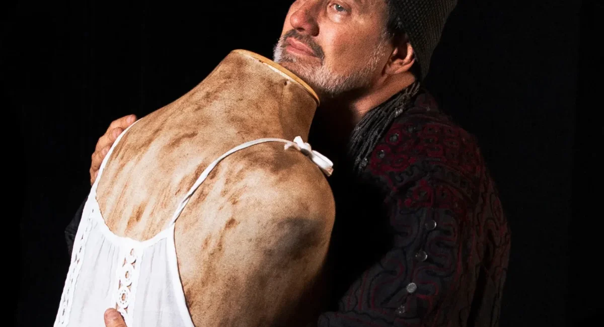 Foto de divulgação de "A Raiz". Claudio Fontana, vestindo um casacão e gorro preto, abraça um manequim sem cabeça. O manequim veste um vestido de renda branco. Ambas as figuras parecem sujas de terra.