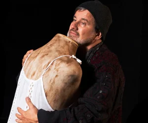 Foto de divulgação de "A Raiz". Claudio Fontana, vestindo um casacão e gorro preto, abraça um manequim sem cabeça. O manequim veste um vestido de renda branco. Ambas as figuras parecem sujas de terra.