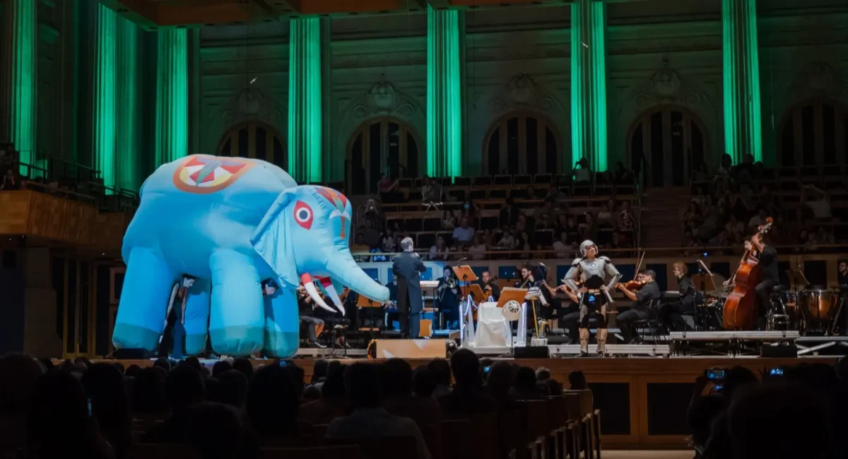 Cena de "Aprendiz de Feiticeiro", encenada pela TUCCA. A Sinfonieta TUCCA Fortíssima está no palco da Sala São Paulo. A frente dela, um fantoche de Elefante, feito de materiais infláveis e um rapaz vestido com armadura