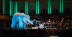 Cena de "Aprendiz de Feiticeiro", encenada pela TUCCA. A Sinfonieta TUCCA Fortíssima está no palco da Sala São Paulo. A frente dela, um fantoche de Elefante, feito de materiais infláveis e um rapaz vestido com armadura