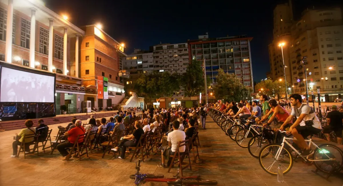 Sessão ao ar livre do BikeCine, realizada em uma praça. Nas primeiras fileiras, cadeiras, ao fundo: pessoas pedalam bicicletas que geram energia para o cinema