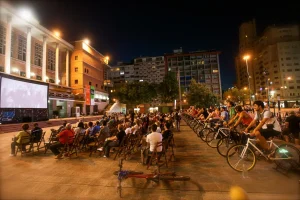 Sessão ao ar livre do BikeCine, realizada em uma praça. Nas primeiras fileiras, cadeiras, ao fundo: pessoas pedalam bicicletas que geram energia para o cinema