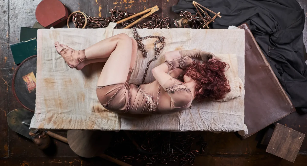 Foto de divulgação "Criatura, uma Autópsia". A atriz Bruna Longo está deitada em posição fetal numa cama. Sua roupa, parecem trapos, que remetem à pele fragmentada de Frankenstein. Ao redor da cama, instrumentos científicos e de embalsamento.