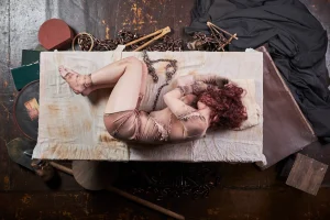 Foto de divulgação "Criatura, uma Autópsia". A atriz Bruna Longo está deitada em posição fetal numa cama. Sua roupa, parecem trapos, que remetem à pele fragmentada de Frankenstein. Ao redor da cama, instrumentos científicos e de embalsamento.