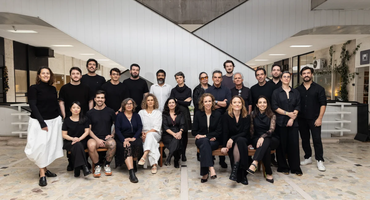 Participantes do 8º Design na Metrópole. Homens e mulheres vestidos de preto (apenas dois com roupa branca) posam para uma foto coletiva. Ao fundo, as escadas rolantes da Galeria Metrópole