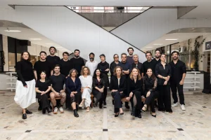 Participantes do 8º Design na Metrópole. Homens e mulheres vestidos de preto (apenas dois com roupa branca) posam para uma foto coletiva. Ao fundo, as escadas rolantes da Galeria Metrópole
