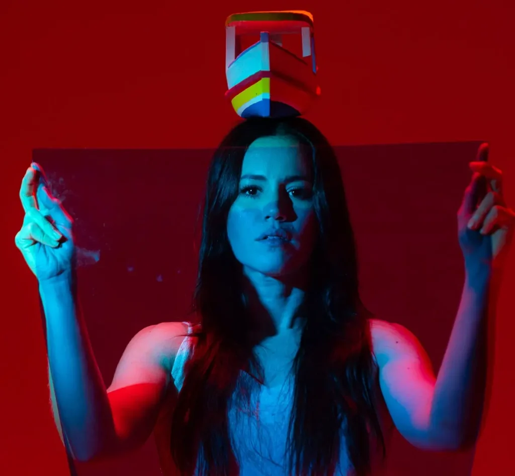 Foto de divulgação de "Do Ventre à Cabeça". Luiza Braga levanta seus braços em gesto de louvação. Um barquinho pousa sobre sua cabeça. A luz tinge sua pele de azul, o fundo é vermelho