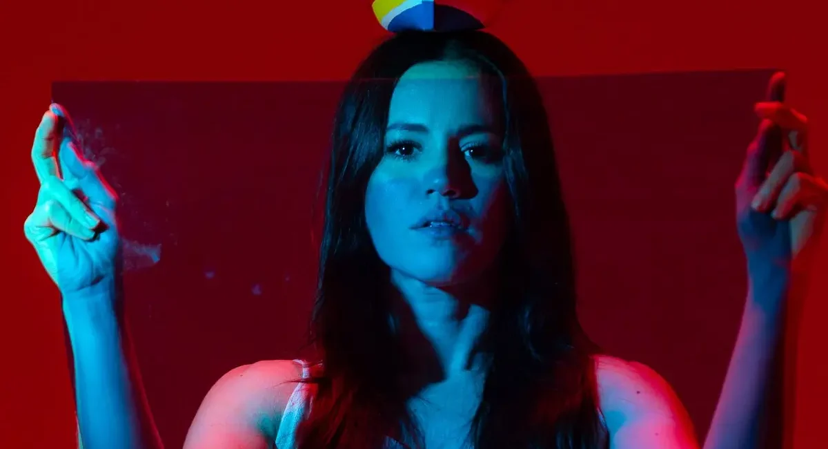 Foto de divulgação de "Do Ventre à Cabeça". Luiza Braga levanta seus braços em gesto de louvação. Um barquinho pousa sobre sua cabeça. A luz tinge sua pele de azul, o fundo é vermelho
