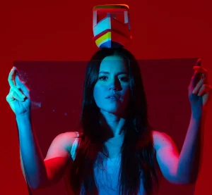 Foto de divulgação de "Do Ventre à Cabeça". Luiza Braga levanta seus braços em gesto de louvação. Um barquinho pousa sobre sua cabeça. A luz tinge sua pele de azul, o fundo é vermelho