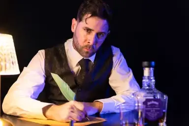 Foto de divulgação de "Edgar Allan Poe Speakeasy". Um ator vestindo camisa, gravata e colete está sentado à mesa empunhando uma caneta de pena. Uma garrafa de gin está colocada na mesa