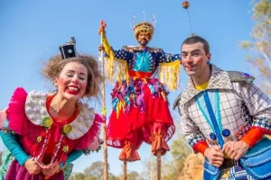 O Núcleo Ás de Paus apresenta "Encontro de Gigantes". Uma palhaça e um palhaço estão agachados olhando para a câmera; ao fundo, outro palhaço em pernas-de-pau também sorri para a câmera. O fundo é o céu azul