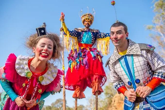 O Núcleo Ás de Paus apresenta "Encontro de Gigantes". Uma palhaça e um palhaço estão agachados olhando para a câmera; ao fundo, outro palhaço em pernas-de-pau também sorri para a câmera. O fundo é o céu azul