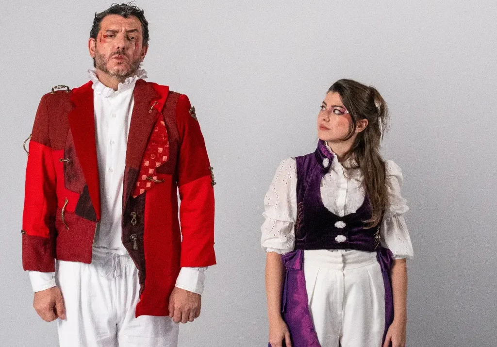 Foto de divulgação do espetáculo. Diego Rodda veste uma casaca vermelha e Luiza Porto um colete roxo-escuro.
