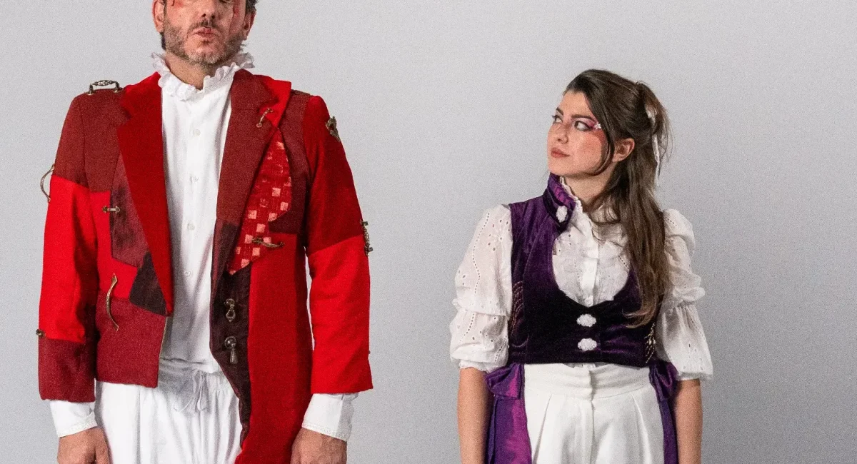Foto de divulgação do espetáculo. Diego Rodda veste uma casaca vermelha e Luiza Porto um colete roxo-escuro.