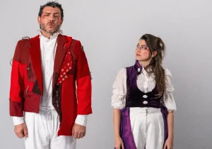 Foto de divulgação do espetáculo. Diego Rodda veste uma casaca vermelha e Luiza Porto um colete roxo-escuro.