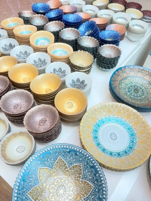 Tigelas e pratos de vidro decorados com mandalas e arabescos em azul, dourado, prata e rosé, empilhados sobre uma bancada da Expo Arabia