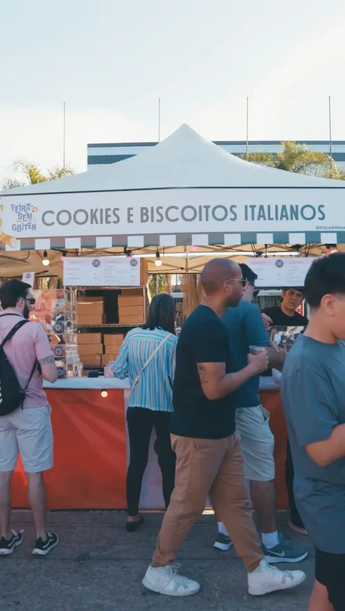 Pessoas fazem fila e circulam diante da barraca "Cookies e Biscoitos Italianos", da Feira Sem Glúten, montada ao ar livre em um dia de sol