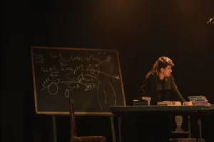Cena de "Galileu". À esquerda, uma lousa recheada de fórmulas astronômicas; à direita, o personagem titular se debruça sobre a mesa de estudos