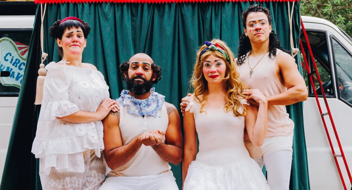 A Trupe DuNavô apresenta o "Grancirco DuNavô". Vestidos de palhaço, eles posam para a foto, possuem vestes brancas e bufantes. O fundo é uma cortina esverdeada.