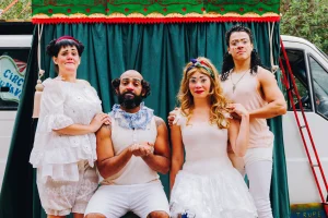 A Trupe DuNavô apresenta o "Grancirco DuNavô". Vestidos de palhaço, eles posam para a foto, possuem vestes brancas e bufantes. O fundo é uma cortina esverdeada.