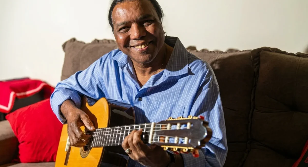 O compositor e cantor Lula Barbosa. Sentado ao sofá, segurando seu violão. Veste uma camisa azul. Sorri para a câmera