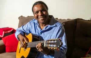 O compositor e cantor Lula Barbosa. Sentado ao sofá, segurando seu violão. Veste uma camisa azul. Sorri para a câmera