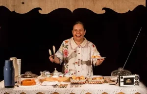 Geni Cavalcante interpreta "O Céu de Durvalina". Em cena, a atriz está atrás de uma grande mesa recheada de bolos e pão-de-queijo, xícaras de café e um radinho. Ela veste camisa branca florida e seu cabelo está preso em um coque. Ela sorri enquanto empunha uma pinça para servir seus fregueses (fora de quadro)