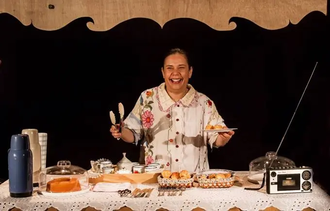Geni Cavalcante interpreta "O Céu de Durvalina". Em cena, a atriz está atrás de uma grande mesa recheada de bolos e pão-de-queijo, xícaras de café e um radinho. Ela veste camisa branca florida e seu cabelo está preso em um coque. Ela sorri enquanto empunha uma pinça para servir seus fregueses (fora de quadro)