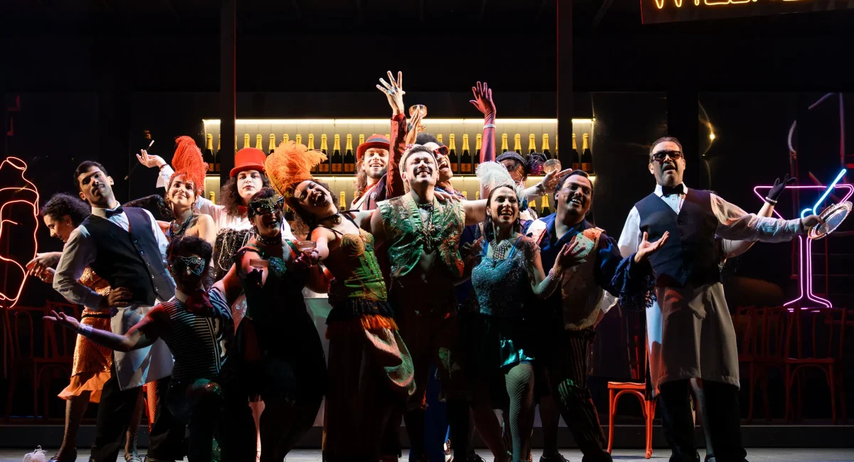 Cena de "O Morcego", encenada pela Academia de Ópera do Theatro São Pedro. Um palco colorido, banhado em luzes neon, com um bar ao fundo, o coro (vestindo uma gala colorida e máscaras) é iluminado enquanto canta.
