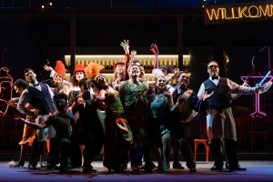 Cena de "O Morcego", encenada pela Academia de Ópera do Theatro São Pedro. Um palco colorido, banhado em luzes neon, com um bar ao fundo, o coro (vestindo uma gala colorida e máscaras) é iluminado enquanto canta.