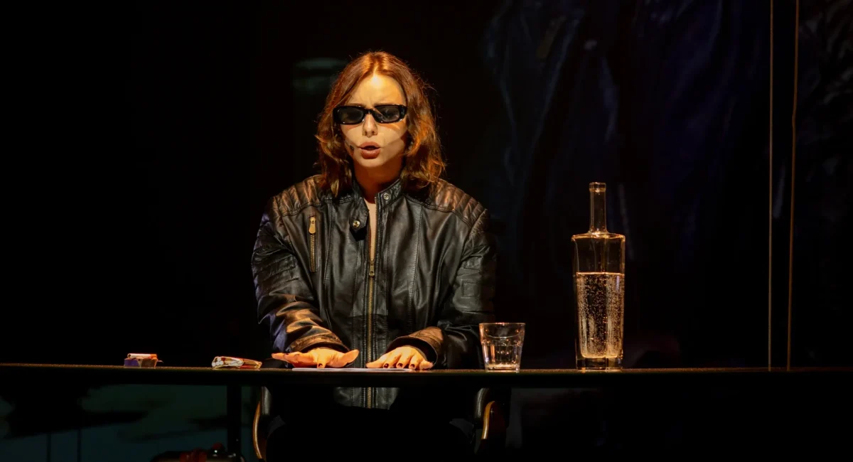 Cena de "O Ovo e a Galinha". Ana Hartmann, a atriz, está sentada à mesa vestindo uma jaqueta preta e óculos escuros. Ela fala em um microfone de mesa, como quem profere uma fala em um simpósio.
