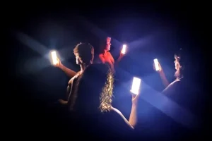 Cena de "QR Corpo", da Anti Status Quo. Em um palco escuro, bailarinos empunham smartphones cujas telas brilhantes iluminam a cena e seus rostos.