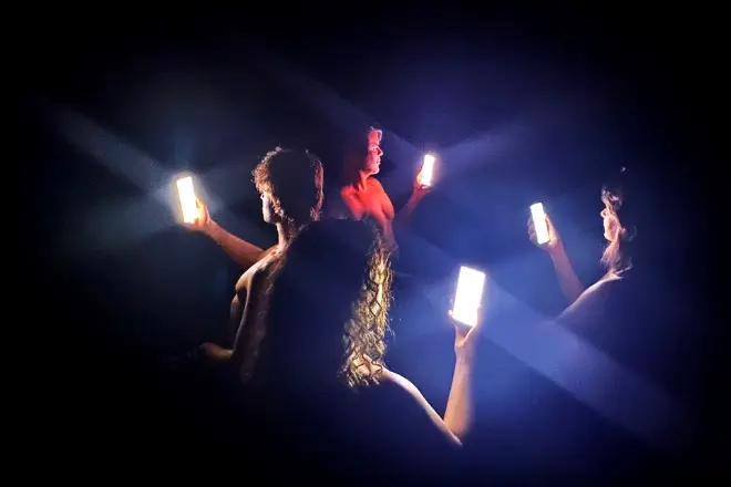 Cena de "QR Corpo", da Anti Status Quo. Em um palco escuro, bailarinos empunham smartphones cujas telas brilhantes iluminam a cena e seus rostos.