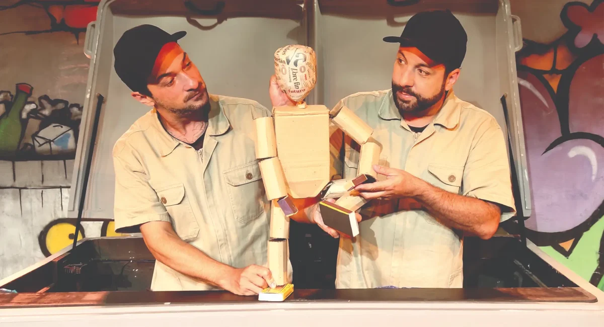 Cena de "Quanta Coisa o Lixo É". Dois atores vestidos de operários manipulam um boneco humanoide feito de papelão