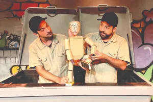 Cena de "Quanta Coisa o Lixo É". Dois atores vestidos de operários manipulam um boneco humanoide feito de papelão