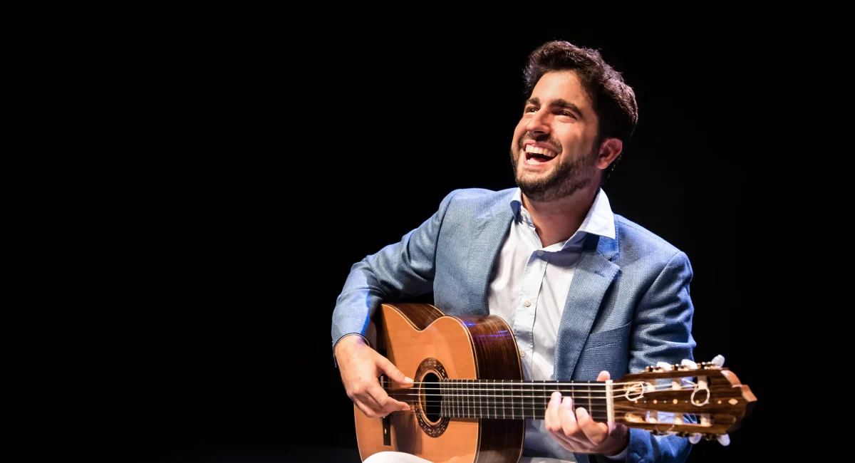 O violonista Rafael Aguirre em apresentação. Ele sorri enquanto toca o violão, veste camisa branca e paletó azul claro