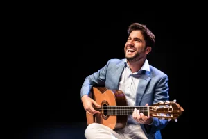 O violonista Rafael Aguirre em apresentação. Ele sorri enquanto toca o violão, veste camisa branca e paletó azul claro