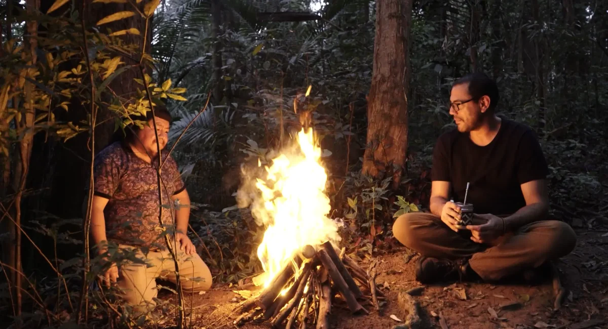 Edgar Calel e Fernando Pereira dos Santos no vídeo "Ru Q'an Ramaj – Escalera del Tiempo". Em uma floresta ao fim do dia, os dois homens estão sentados um de cada lado de uma fogueira que ilumina seus corpos.