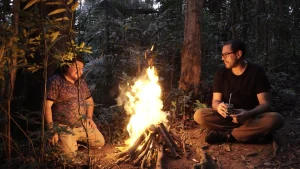 Edgar Calel e Fernando Pereira dos Santos no vídeo "Ru Q'an Ramaj – Escalera del Tiempo". Em uma floresta ao fim do dia, os dois homens estão sentados um de cada lado de uma fogueira que ilumina seus corpos.