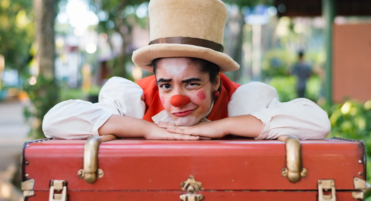Fernanda Nunes em "Tertulia e o Fantástico Circo de Pulgas". Vestida de palhaço, com uma cartola branca, ela se debruça sobre uma mala. Está ao ar livre, uma praça com árvores desfocada ao fundo