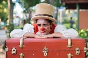 Fernanda Nunes em "Tertulia e o Fantástico Circo de Pulgas". Vestida de palhaço, com uma cartola branca, ela se debruça sobre uma mala. Está ao ar livre, uma praça com árvores desfocada ao fundo