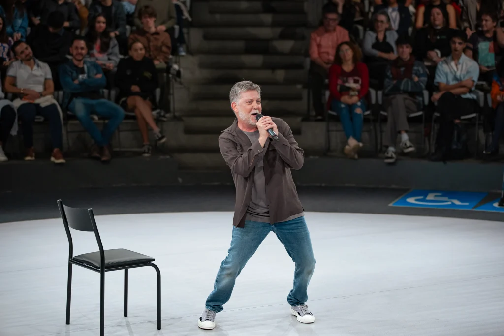 Kiko Mascarenhas interpreta "Todas as Coisas Maravilhosas". O ator segura um microfone e canta no centro do palco circular do Tucarena. Ao fundo, a plateia observa a apresentação