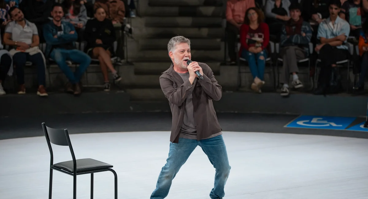 Kiko Mascarenhas interpreta "Todas as Coisas Maravilhosas". O ator segura um microfone e canta no centro do palco circular do Tucarena. Ao fundo, a plateia observa a apresentação